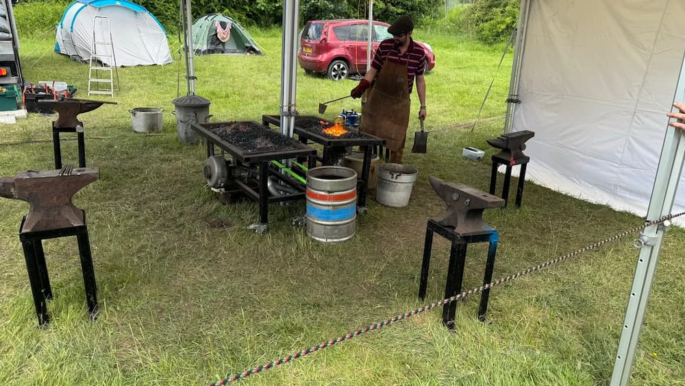 A white tent covering 4 anvils and two smelting stations. A man in a leather apron is using some tools to interact with the hot coals