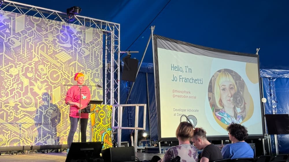 A photo of Jo Franchetti stood on a stage behind a podium with their presentation showing on a screen to the right